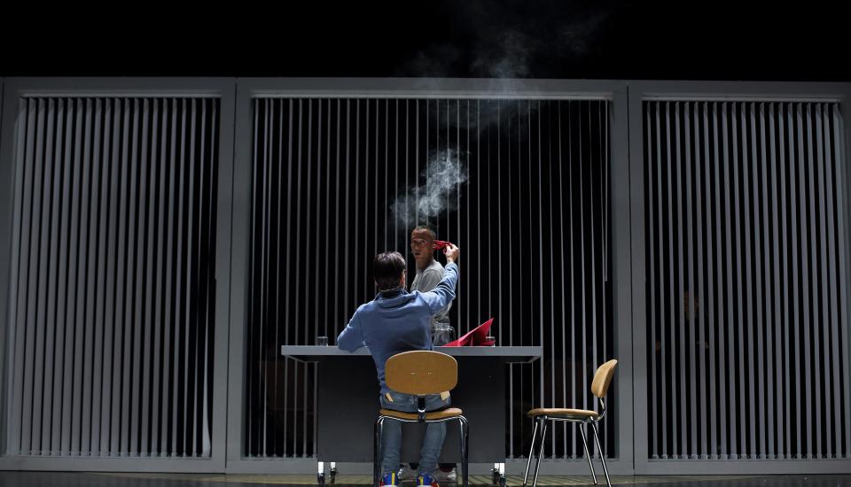 Une personne asssise à son bureau, vue de dos, semble brûler un morceau de papier rouge. Une autre personne, en face du bureau, regarde. 
