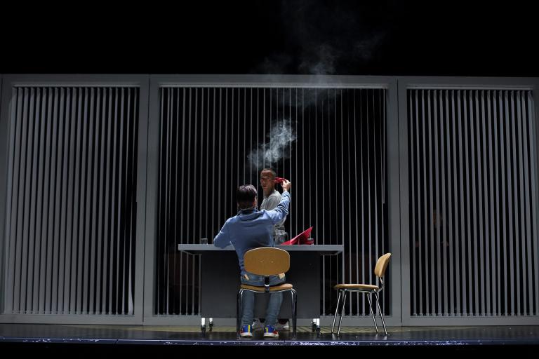 Une personne asssise à son bureau, vue de dos, semble brûler un morceau de papier rouge. Une autre personne, en face du bureau, regarde. 