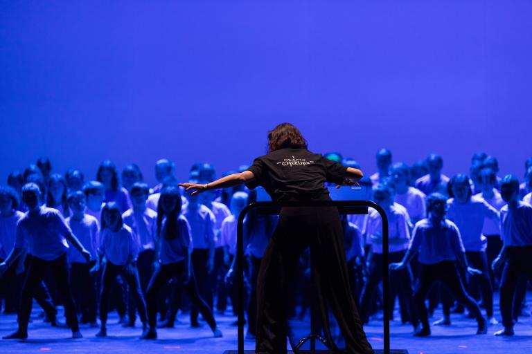 Une cheffe de chœur vue de dos, animant un chœur d'enfant. 
